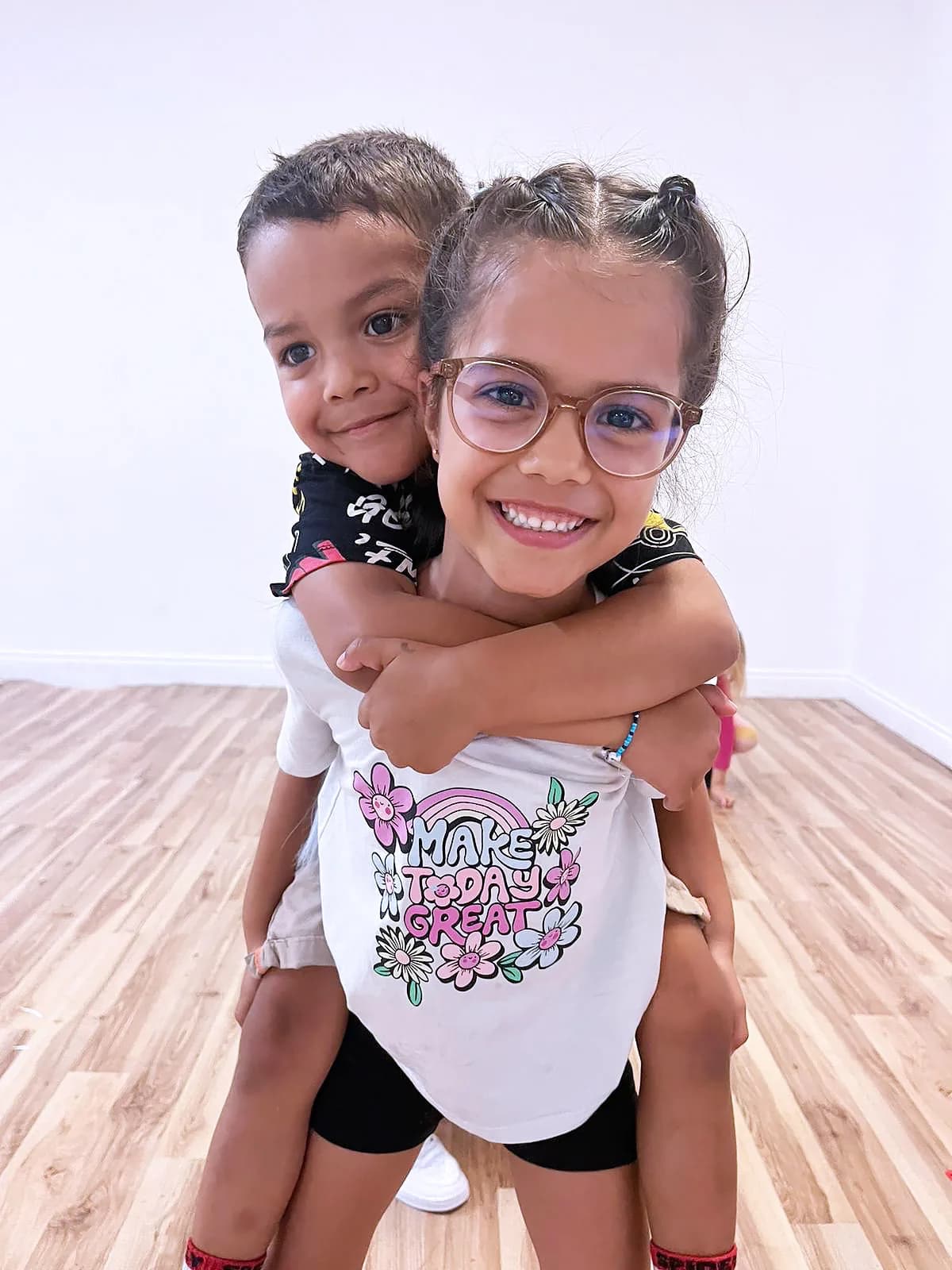 Two HK Minis dancers in the studio, one piggybacking the other, both grinning in matching pose.